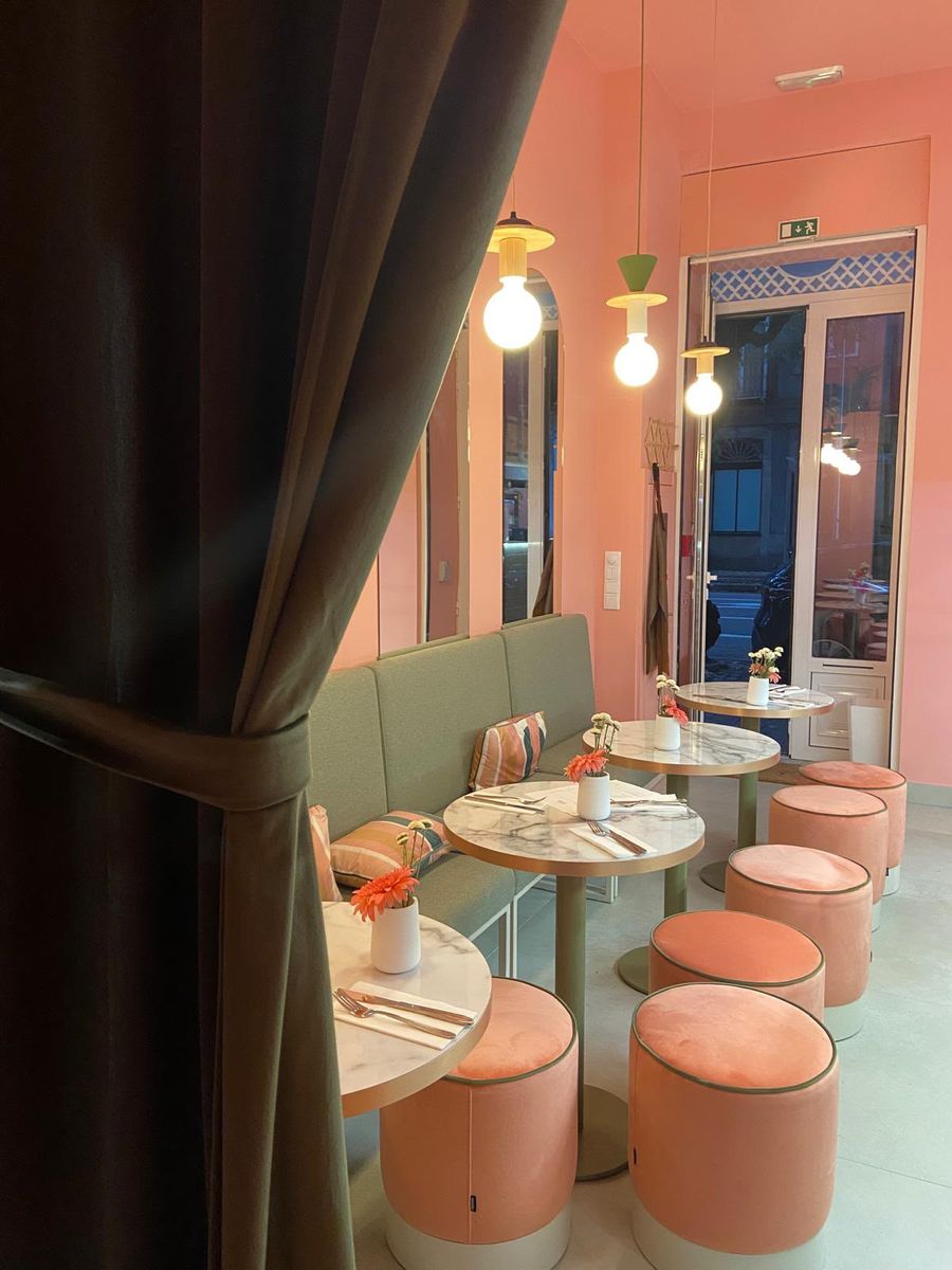 The dining room at Hora Mansa: salmon-pink walls, a sage-green banquette, round marble tables with fresh flowers, peach velvet stools, pendant lights and an olive curtain, with the glass door opening onto the street in Bonfim, Porto. / A sala da Hora Mansa: paredes rosa-salmão, banco corrido verde-sálvia, mesas redondas de mármore com flores frescas, bancos de veludo pêssego, candeeiros suspensos e um cortinado verde-azeitona, com a porta envidraçada para a rua, no Bonfim, Porto.