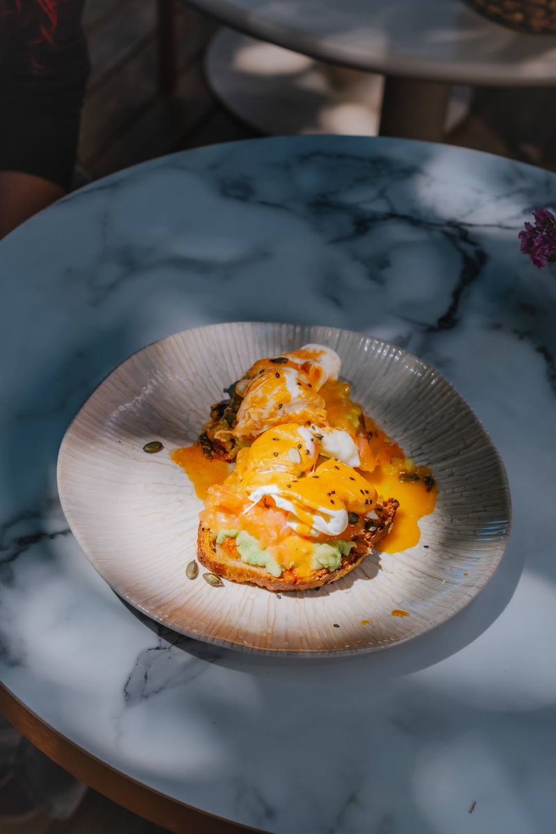 Nordic Benny — poached eggs draped in hollandaise over smoked salmon and smashed avocado on artisan toast, sprinkled with seeds, served on a marble table. / Nordic Benny — ovos escalfados cobertos de molho holandês sobre salmão fumado e abacate esmagado em tosta artesanal, com sementes, servido numa mesa de mármore.